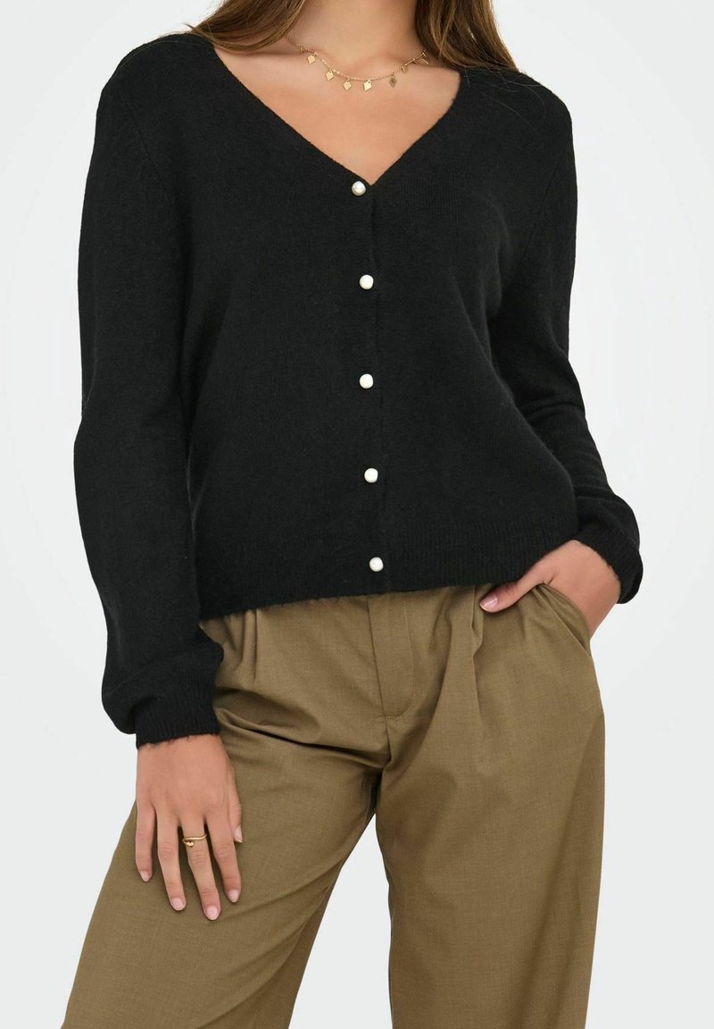 Femme portant un cardigan noir col en V avec des boutons en perle et un pantalon plissé taille haute olive, main dans la poche, collier en or et bague.