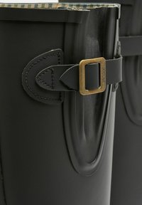 Črne gumijaste dežne škornje z teksturirano površino. Opremljene so z usnjenim trakom in bakrenim sponko, kar poudarja tradicionalen dizajn.