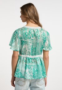 Blusa verde con motivo floreale, maniche corte a sbuffo, bordo in pizzo sul collo e vita a peplo. Indossata con jeans in denim chiaro.