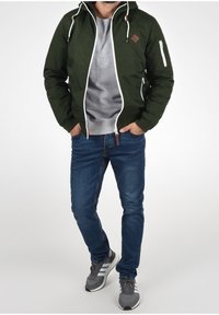 Giacca verde con zip e cappuccio, dettagli bianchi, logo a rombo e tasche laterali, abbinata a una felpa grigia e jeans blu.