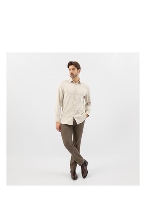 Uomo con camicia beige a bottoni, pantaloni marroni e stivali marrone scuro, in piedi con una mano in tasca e le gambe incrociate su sfondo bianco.