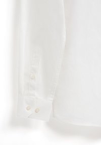 Chemise blanche à manches longues avec un poignet pointu, comportant deux fermetures à bouton et une texture de coton lisse. Design épuré sans motifs.