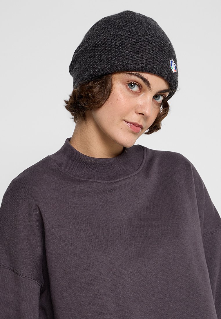 Młoda osoba z krótkimi kręconymi włosami, nosząca teksturowaną czarną ciepłą czapkę i szary sweter z wysokim kołnierzem, zwrócona lekko bokiem.
