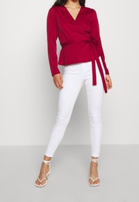 Blouse cache-cœur bordeaux à manches longues avec ceinture à la taille, associée à un jean skinny blanc et des sandales blanches à lanières. Tissu lisse et coupe ajustée.