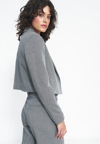 Szara krótka kurtka w wzór houndstooth, z długimi rękawami i dopasowanym krojem, noszona z pasującymi spodniami.