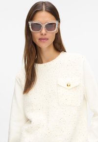 Priehľadné mačacie okuliare so svetlohnedými tónovanými šošovkami, skombinované s texturovaným bielym svetríkom s detailom zlatej gombíkovej kapsy.