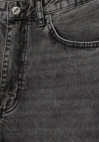 Gros plan sur un jean en denim noir montrant la poche avant, la boucle de ceinture et le bouton métallique avec le logo de la marque gravé.