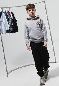Sudadera gris con logo, pantalones cargo negros y zapatillas gruesas. Estante de exhibición con varias camisetas de fondo. Texturas suaves.