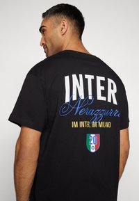 Svart T-skjorte med "INTER" i fete hvite bokstaver, "Nerazzurri" i blå håndskrift, og gulltekst. Inkluderer et italiensk flaggmerke med nummer 20.