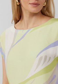 Hellgrüne Bluse mit abstrakten Mustern in Lavendel und Beige, Rundhalsausschnitt und glatter, weicher Stoffstruktur. Goldene Creolen.