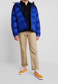 Blauwe gewatteerde jas met zwarte accenten, ritsclosure en capuchon, gedragen over een zwart button-up shirt. Beige broek en oranje sneakers maken de outfit compleet.