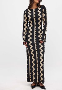 Robe noire longue avec un motif en zigzag beige, fabriquée à partir d'un tissu texturé, avec des manches longues et une silhouette ajustée. Des chaussures noires complètent la tenue.