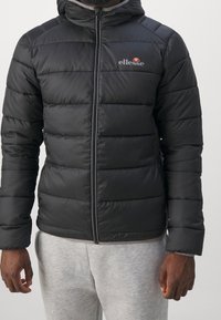 Czarna pikowana kurtka z kapturem. Posiada poziome przeszycia, zamek z przodu oraz logo na lewej piersi. Materiał wydaje się błyszczący.