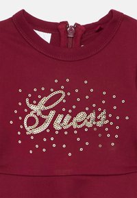 Camisa bordô com um fecho na parte de trás e um logotipo "Guess" em lantejoulas douradas, com lantejoulas espalhadas por todo o tecido.