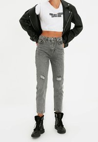 Veste en cuir noir de style motard sur un t-shirt court blanc avec un texte graphique. Porté avec un jean gris déchiré taille haute et des bottes de combat noires.