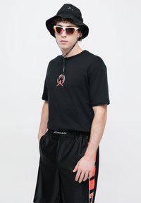 Μαύρο βαμβακερό T-shirt με κόκκινο λογότυπο, συνδυασμένο με μαύρο αθλητικό σορτς με κόκκινες λεπτομέρειες. Φορεμένο με μαύρο καπέλο τύπου bucket και γυαλιά ηλίου.