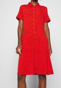 Červené šaty s krátkým rukávem a zapínáním na knoflíky vpředu, s černým skvrnitým vzorem. Rozšířená sukně a límečkový design. Vyrobeno z lehkého materiálu.