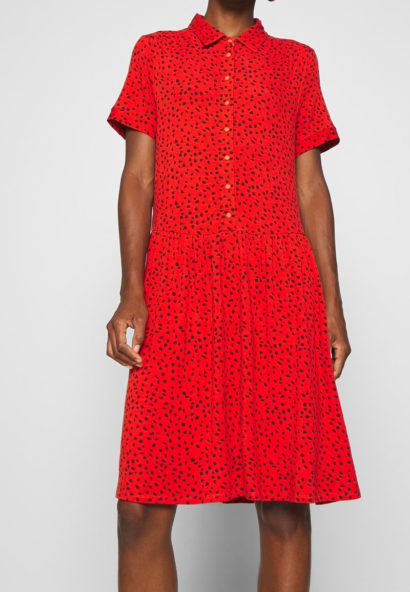 Červené šaty s krátkým rukávem a zapínáním na knoflíky vpředu, s černým skvrnitým vzorem. Rozšířená sukně a límečkový design. Vyrobeno z lehkého materiálu.