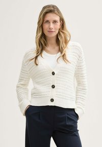 Weißer Strickcardigan mit offenem Gewebe-Muster, gerippten Bündchen und fünf großen schwarzen Knöpfen. Kombiniert mit einem weißen Oberteil und navyblauen Hosen.