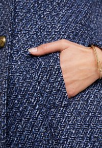 Hand med guldarmband i en blå och vit texturerad kappa, som visar en närbild av tyg och knappdetaljer.
