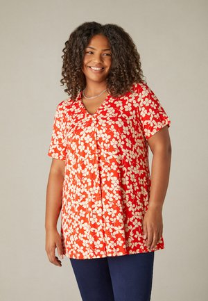 Blusa rossa a fiori con maniche corte, caratterizzata da un scollo a V e motivi floreali bianchi, realizzata in tessuto leggero. Abbinata a pantaloni blu.