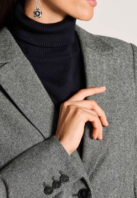 Grijze houndstooth blazer met notch-lapels, zwarte knopen, gecombineerd met een zwarte coltrui. Close-up van een hand die de kraag van de blazer aanpast.