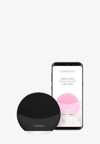 Svart silikon ansiktsrensingsenhet med teksturert overflate, ved siden av en smarttelefon som viser et oppsett skjermbilde med en rosa versjon av enheten.