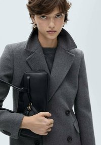 Gray wool-blend coat with a double-breasted design, worn over a dark sweater. Black leather handbag with a shoulder strap is held in one hand.