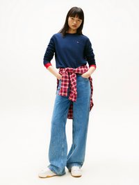 Sweatshirt marine à col rond avec des accents rouges, associé à un jean denim bleu à jambes larges. Une chemise écossaise rouge est nouée autour de la taille. Baskets blanches.