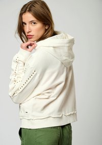 Sudadera blanca con detalles de tachuelas doradas a lo largo de las mangas, puños acanalados y un dobladillo sin acabar. Cuenta con una capucha ajustable y una textura de tela suave.