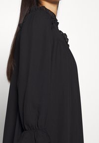 Blouse noire avec un col haut et des volants, des manches longues avec des poignets élastiques, et une texture de tissu lisse et fluide.