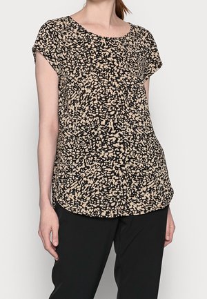 Kobieta ubrana w beżową bluzkę z krótkim rękawem z abstrakcyjnym czarnym wzorem i czarne spodnie, stojąca na jednolitym tle.