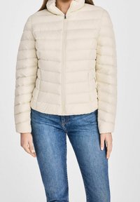 Kobieta w kremowej pikowanej kurtce puchowej z kołnierzem stojącym i zapięciem na zamek z przodu, w połączeniu z niebieskimi dżinsami, na jednolitym tle.