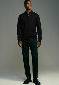 Schwarzes Poloshirt mit drei Knöpfen, kombiniert mit dunkelgrünen Hosen. Das Outfit wird durch schwarze Stiefeletten ergänzt. Schlichtes und modernes Design.