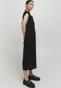 Abito lungo nero senza maniche con fessure laterali; tessuto strutturato; scollatura rotonda; abbinato a mocassini neri spessi con accenti dorati.