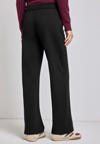 Zwarte wijde broek heeft een gladde textuur, een elastische tailleband en een schoon ontwerp zonder zichtbare sluitingen. Gecombineerd met beige schoenen.