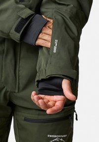 Zielona oliwkowa kurtka wodoodporna z regulowanymi mankietami, czarną podszewką i logo. Posiada praktyczny design z wzmocnionymi szwami.