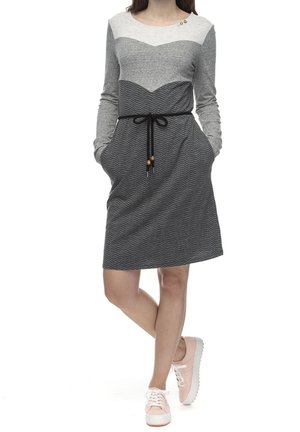 Kobieta ubrana w długą, sięgającą kolan, szarą sukienkę w wzory z czarnym paskiem w talii, stojąca z rękami w kieszeniach, w różowych trampkach.