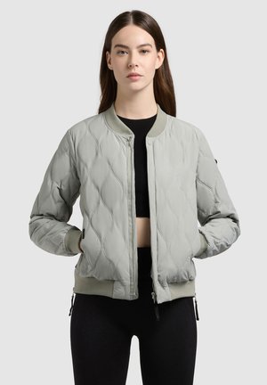 Moteris dėvi šviesiai pilką su kuprelėmis bomberio striukę ant juodo apkarpyto viršaus ir juodų kelnių, stovinti su rankomis kišenėse.