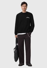 Felpa nera con logo "AllSaints" bianco, indossata sopra una camicia bianca, abbinata a pantaloni scuri con accento a catena e con una borsa nera in mano.