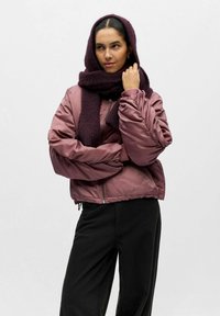 Blouson aviateur matelassé en satin rose poussiéreux, avec fermeture éclair et poignets côtelés, associé à une écharpe texturée violet foncé. Pantalon noir.