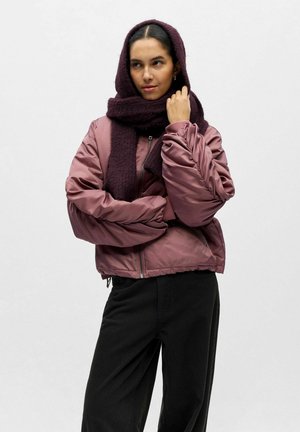 Blouson aviateur matelassé en satin rose poussiéreux, avec fermeture éclair et poignets côtelés, associé à une écharpe texturée violet foncé. Pantalon noir.