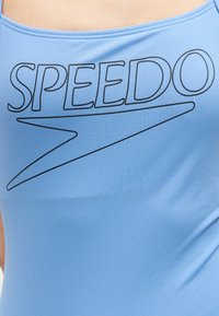 Gros plan sur un tissu bleu clair avec le logo « SPEEDO » et un motif swoosh imprimés en noir au niveau de la poitrine.