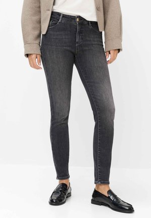 Personne portant un jean slim gris foncé, une veste beige, un haut blanc et des mocassins en cuir noirs, debout devant un fond blanc uni.