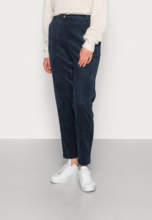Person trägt marineblaue Cordhose, weißen Pullover und weiße Turnschuhe und steht auf einem hellgrauen Boden vor einer weißen Wand.