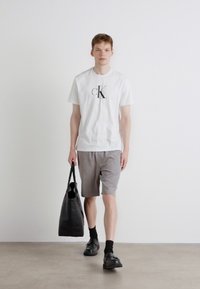 Balta medvilnės marškinėliai su juodu "cK" logotipu, derinami su pilkais šortais ir juodais batais. Modelis laiko juodą rankinę. Minimalistinis fonas.