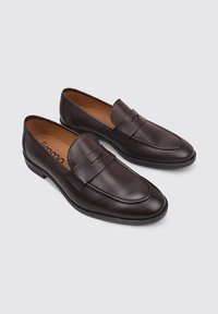 Mocasines de piel marrón con una textura suave, que presentan un sutil diseño de costura, una correa rectangular sobre la parte delantera y un tacón bajo.