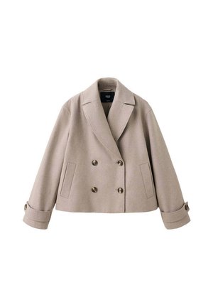 Giovane donna in piedi che indossa un cappotto doppiopetto beige, maglione grigio, jeans blu a gamba larga e scarpe nere su uno sfondo beige uniforme.