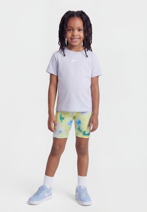 Κορίτσι που στέκεται, φορώντας ένα ελαφρύ μωβ μπλουζάκι Nike, κίτρινα σορτς με λουλούδια, λευκές κάλτσες και ανοιχτό μπλε αθλητικά παπούτσια Nike σε λευκό φόντο.