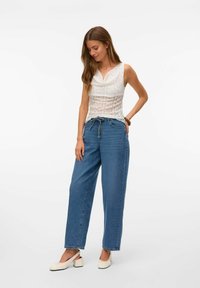 Haut sans manches blanc avec une texture plissée, associé à un jean en denim bleu ample et des chaussures à talons blocs crème, se tenant sur un fond uni.